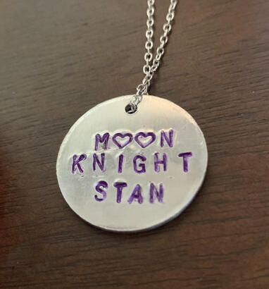 a circular silver pendant on a necklace, featuring the text "moon knight stan" - the o's in moon are replaced with two hearts - the text is inked in purple